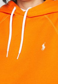 Oranžová mikina s kapucí vyrobená z měkké tkaniny s bílým stahovacím šňůrkou, která má malé bílé logo na prsou. Klasický puloverový design.