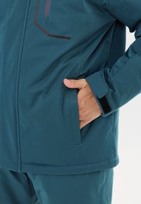 Person, der eine türkisfarbene Jacke und Hose trägt, mit der Hand in der Jackentasche, wobei der Klettverschluss am Ärmelsaum sichtbar ist.