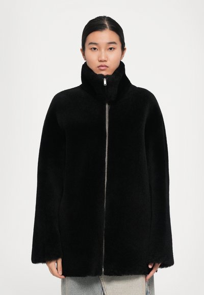 Frau in einer schwarzen, hochkragen Fleecejacke mit einem Reißverschluss vorne, steht vor einem hellen, einfarbigen Hintergrund.