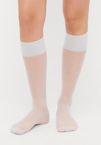 Ponožky po kolena, průhledné bílé barvy s hladkým povrchem a elastickým páskem na vrchu. Mají otevřenou špičku s minimalistickým vzhledem.