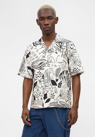 Άνδρας με κοντά ξανθά μαλλιά που φοράει λευκό κοντομάνικο πουκάμισο με μαύρο αφηρημένο και ζωικό μοτίβο, σε συνδυασμό με μπλε τζιν.