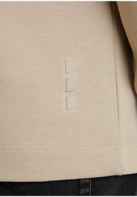 Heller beige, strukturiertes Gewebe mit drei kleinen quadratischen Akzenten in einem helleren Farbton, die vertikal an der Seitennaht angebracht sind.