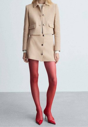 Veste cropped en beige texturé avec deux poches et des boutons argentés, assortie à une mini-jupe et des collants rouges vifs avec des talons.