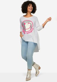 Hellgraune Tunika mit silbernem Smiley-Gesicht und pinkem Text, kombiniert mit blauen Skinny-Jeans und metallischen Stiefeletten.