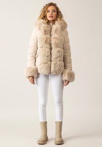 Beige Daunenjacke mit einem langen Kragen und Manschetten aus Kunstfell, ausgestattet mit einem gesteppten Design und einer plüschigen Textur, kombiniert mit weißen, eng anliegenden Hosen.