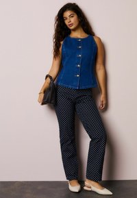 Farmerkék ujjatlan felső arany gombokkal, tengerészkék pöttyös nadrággal párosítva. A modell egy fekete táskát tart, és világos színű lapos cipőt visel.