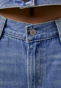 Světle modré džíny s vysokým pasem, s měděným zapínáním na knoflík a viditelnými švy. Texturovaná tkanina s mírně vybledlým povrchem.