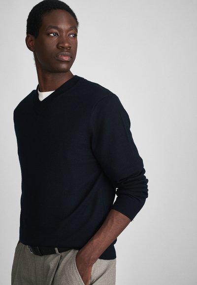 Giovane uomo che indossa un maglione V-collo blu navy sopra una camicia bianca e pantaloni beige, in piedi con una mano in tasca, che guarda di lato.