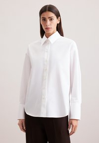 Camicia bianca con colletto a punta, maniche lunghe e orlo leggermente curvato. Realizzata in tessuto liscio con una leggera lucentezza.
