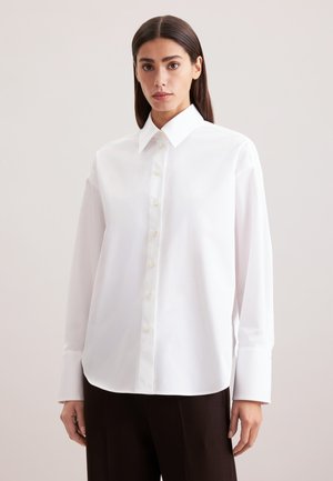 Camicia bianca con colletto a punta, maniche lunghe e orlo leggermente curvato. Realizzata in tessuto liscio con una leggera lucentezza.