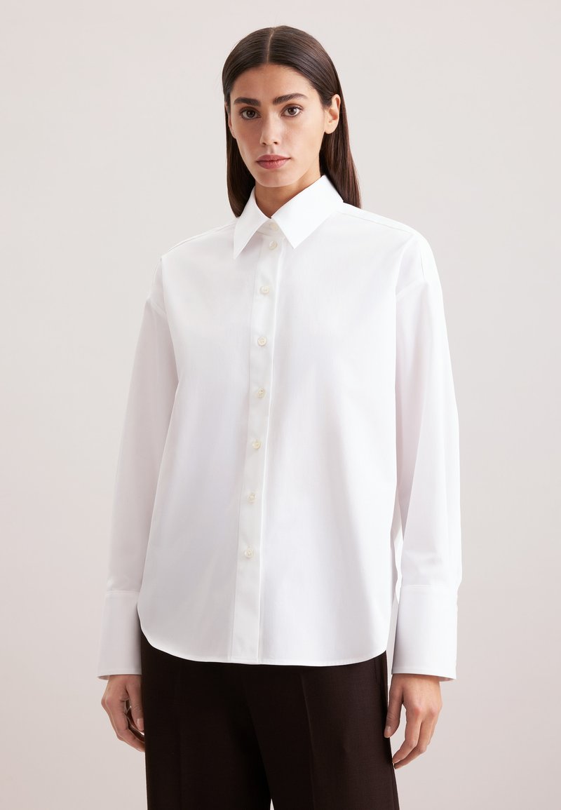 Camicia bianca con colletto a punta, maniche lunghe e orlo leggermente curvato. Realizzata in tessuto liscio con una leggera lucentezza.