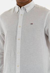 Camicia bianca con bottoni realizzata in un tessuto leggero, con colletto classico, bottoni frontali e un piccolo logo rosso, bianco e blu.