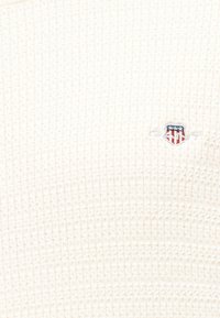 Kremowa dzianina wafelkowa z małym haftowanym logo w kształcie tarczy, przedstawiającym gwiazdy i pasy w kolorach czerwonym, białym i niebieskim.