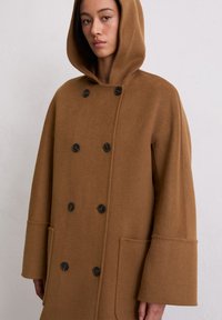 Manteau en laine marron avec capuche, design croisé à double boutonnage, boutons noirs, grandes poches et manches larges. Texture douce avec une finition soignée.