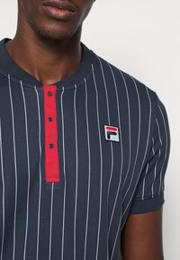 Mörkblå randig polo-skjorta med röd knappslå och broderad logotyp. Har en rund halsringning och korta ärmar med en mjuk textur.