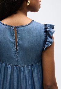 Kobieta o ciemnej karnacji, ubrana w niebieską sukienkę dżinsową z falbaniastymi krótkimi rękawami i zapinanym na guziki wycięciem na plecach, widziana od tyłu i lekko zwrócona.