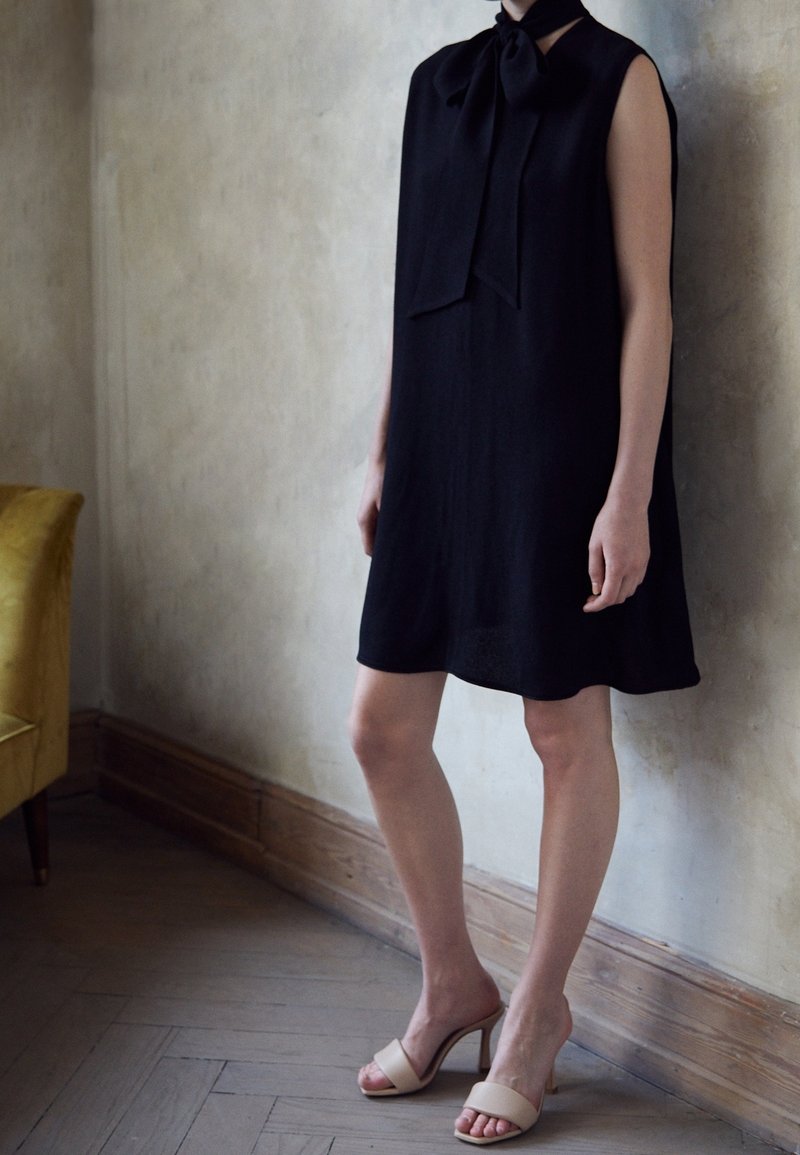 Svart ärmlös klänning med en slipsknut vid halsen, som har en lös, rak passform. Modellen bär beige klacksandaler. Minimalistisk design.