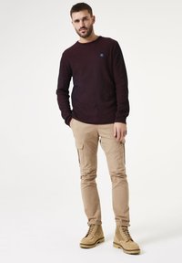 Camisa de manga larga a rayas en rojo oscuro y azul marino con cuello redondo, combinada con pantalones cargo beige y botas de cordones color marrón claro.