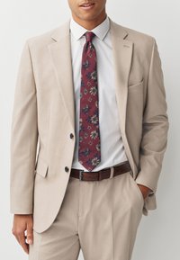 Costume beige en tissu léger, associé à une chemise blanche et une cravate fleurie rouge foncé. Ceinture marron avec une boucle simple.