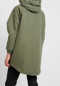 Khaki green parka jacket with a hood, elastic cuffs, and a longer back hem. Made from soft, textured fabric with a smooth finish.