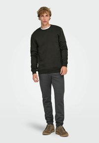 Jeune homme portant un sweat-shirt vert foncé, un pantalon de survêtement gris et des chaussures marron à lacets, debout devant un fond clair uni.