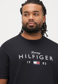 Μαύρο βαμβακερό T-shirt με κοντά μανίκια, με λευκό λογότυπο "Tommy HILFIGER" και το έτος "1985" μαζί με μια ορθογώνια σημαία τριών χρωμάτων.