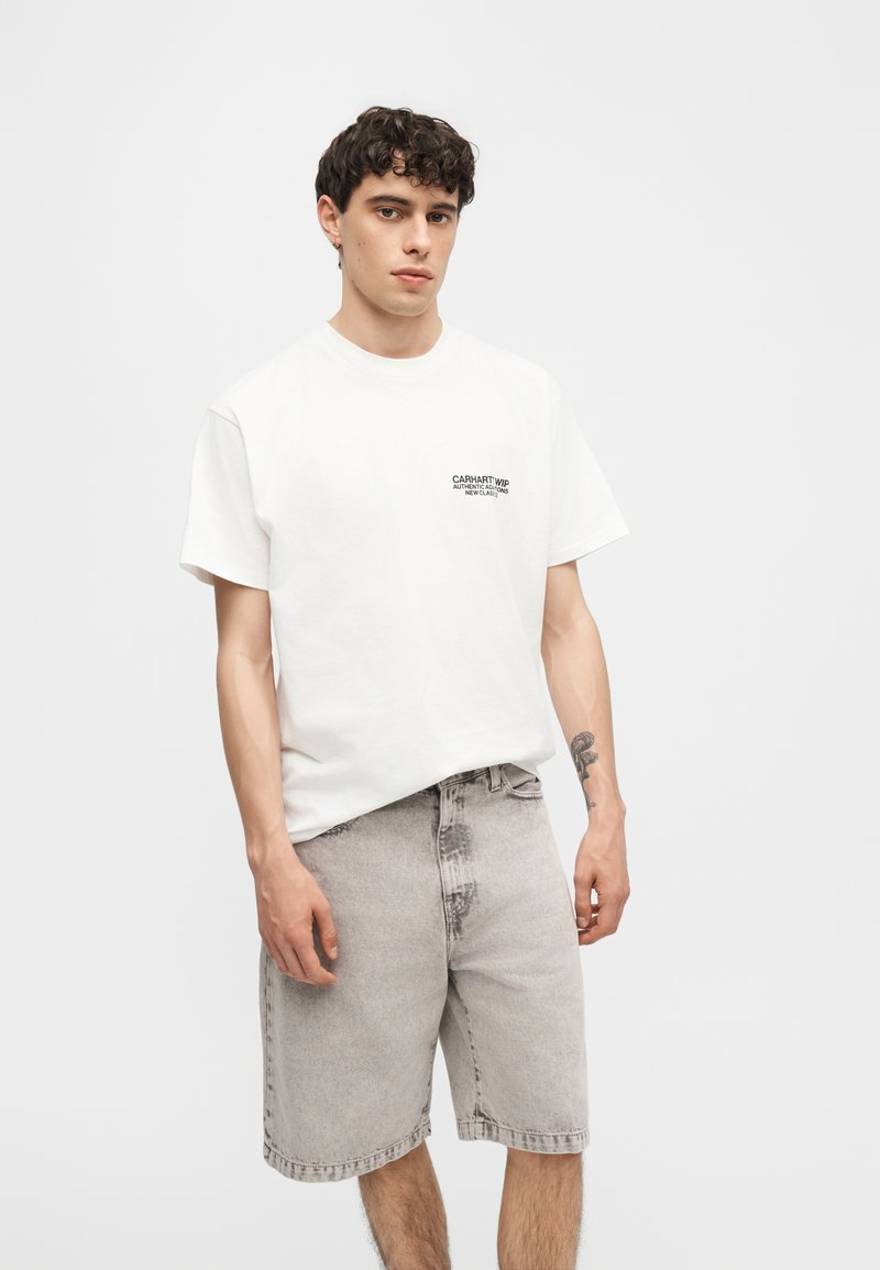 Giovane con capelli corti e ricci che indossa una maglietta bianca Carhartt e pantaloncini di jeans grigio chiaro, in piedi davanti a uno sfondo neutro.