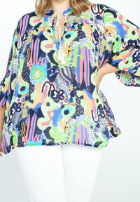 Femme portant un chemisier ample, coloré, à motif abstrait, avec un col à nouer orné de pompons et un pantalon blanc, main reposant sur la hanche.