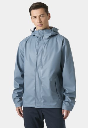 Jeune homme de face portant une veste à capuche imperméable bleu clair avec des poches zippées et un pantalon noir.