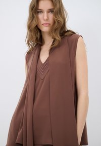 Bruine mouwloze top met een V-hals ontwerp, voorzien van gelaagd detail en een gladde textuur, aangevuld met een lichte drapering.