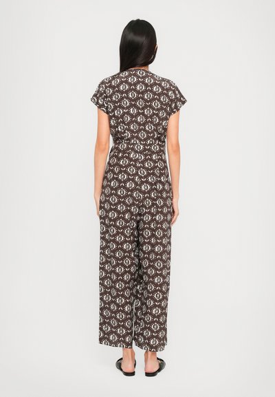 Žena stoji okrenuta leđima prema kameri, nosi smešak jumpsuit u smešnom uzorku s bijelim cvjetovima i crne sandale bez petica, na jednobojnoj pozadini.