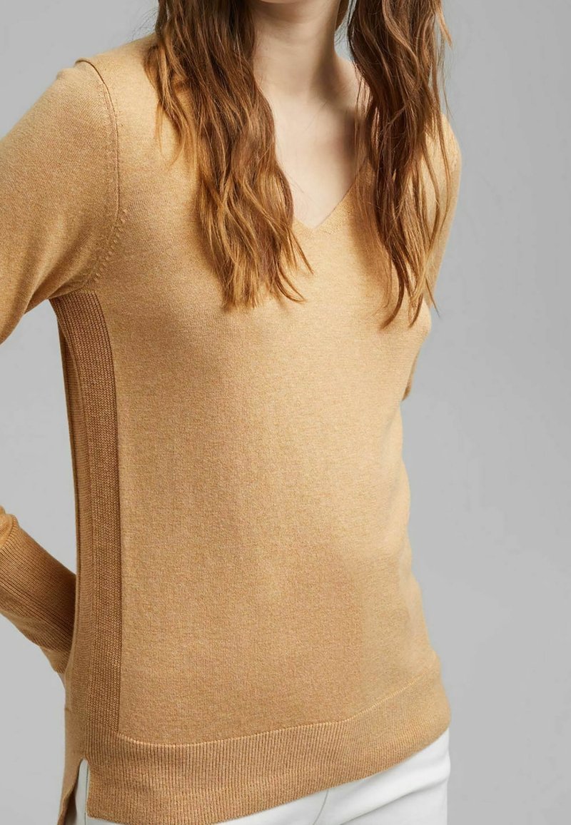 Femme portant un pull beige à col en V avec des panneaux latéraux côtelés et un pantalon blanc sur fond gris uni.