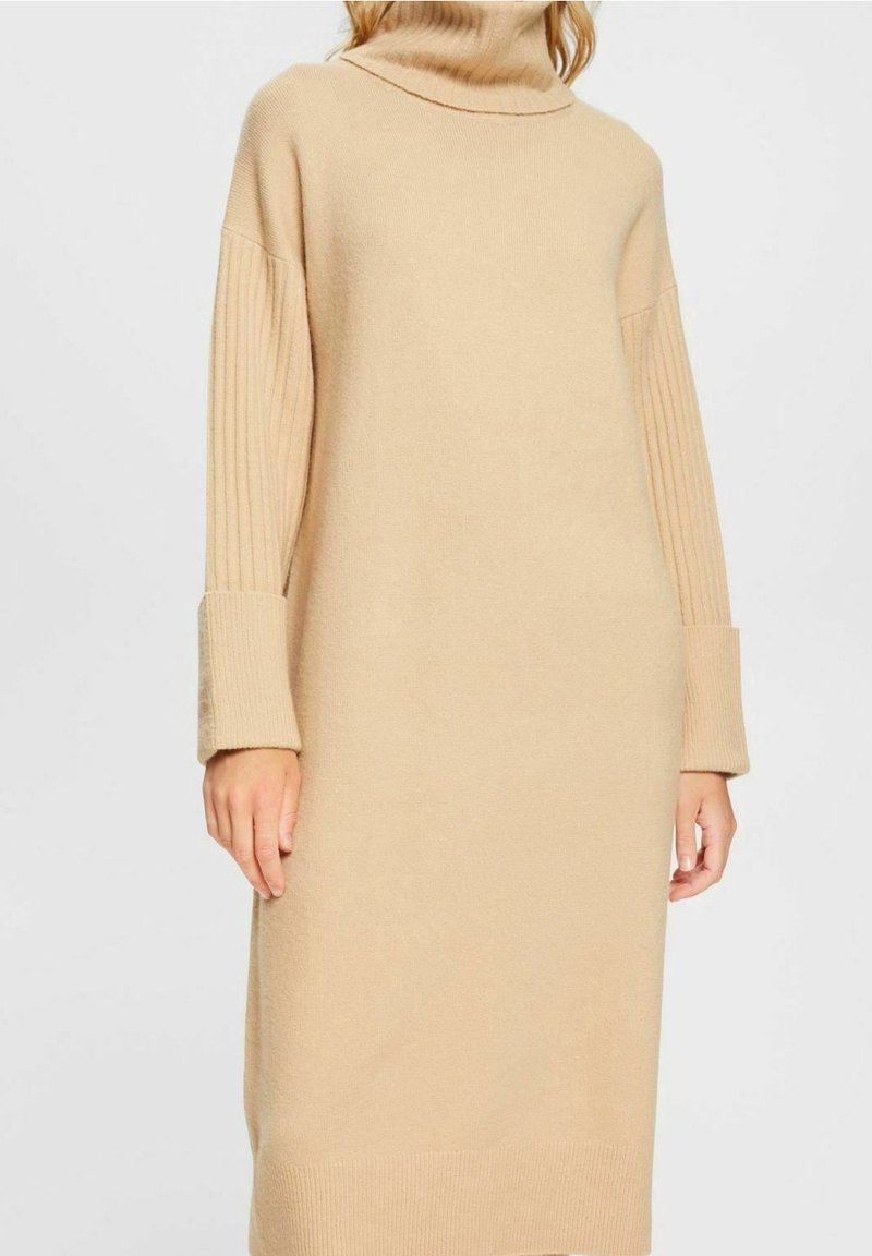 Femme portant une robe-pull beige à manches longues avec des poignets côtelés et un col montant en roulé, sur un fond uni.