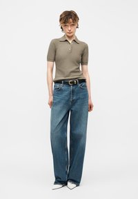 Άτομο που φοράει χακί μπλουζάκι πόλο με κοντά μανίκια, φαρδιά μπλε τζιν παντελόνια με μαύρη ζώνη και λευκά μυτερά παπούτσια, στέκεται μπροστά σε απλό λευκό φόντο.