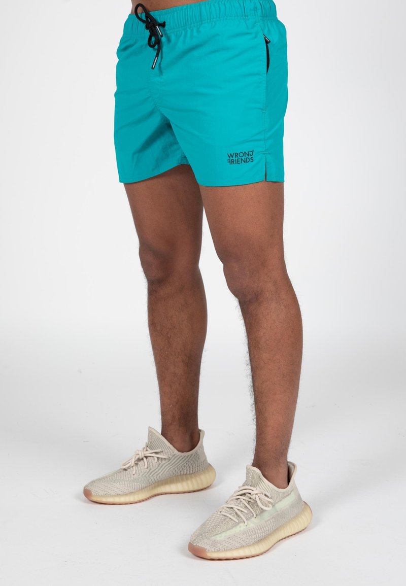 Pantaloni da bagno turchesi con vita regolabile, tasche laterali e dettaglio logo nero. Abbinati a sneaker beige e marroni chiari.