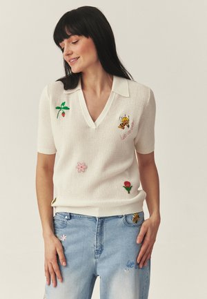 Kobieta w białym sweterkowym polo z haftowaną pszczołą i kwiatami, w połączeniu z jasnoniebieskimi dżinsami ozdobionymi podobnymi naszywkami z kwiatami i pszczołami.