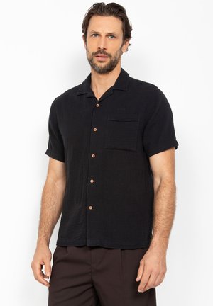 Mand med kort mørkt hår og skæg iført en sort kortærmet skjorte med knapper og mørke bukser, stående foran en hvid baggrund.