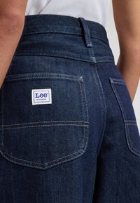 Οπίσθια όψη ατόμου που φοράει σκουρομπλε τζιν Lee Workwear με επιδιορθωτικό έμπλαστρο στην πίσω τσέπη και λευκό τοπ μπουφάν.