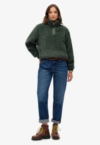 Groene fleecetop met een halve rits en een gestructureerd oppervlak, gecombineerd met blauwe jeans met omslagen en bruine veterschoenen met gele accenten.
