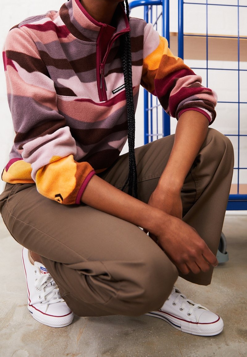 Una persona está agachada vistiendo un pulóver de felpa a rayas en tonos de rosa, marrón y naranja, pantalones caqui y zapatillas Converse blancas.