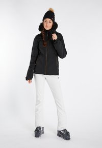 Zwarte geïsoleerde jas met een bont-voering kap, gecombineerd met witte skibroeken en zwarte skibotten. Model heeft een gevlochten haarstijl en een muts.