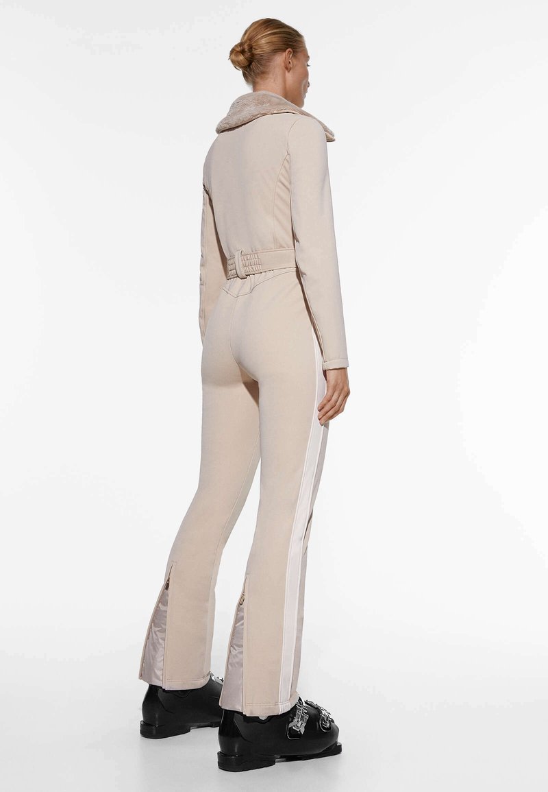 Mono beige con un cuello de pelo, diseño ceñido, piernas acampanadas con cremallera y franjas laterales blancas en contraste. Calzado negro con herrajes decorativos.