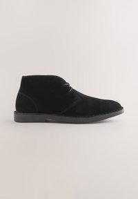 Schwarzer Wildleder-Stiefelette mit zwei Ösen, abgesteppten Details und flacher Gummisohle auf beigem Hintergrund.