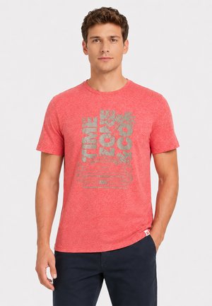 Mann von vorn, trägt ein rotes Grafik-T-Shirt mit dem Text "TIME LOVE COX" und einer Auto-Illustration, dazu eine dunkle Hose vor schlichtem Hintergrund.