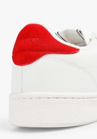 Zapatilla blanca con suela de goma texturizada, cuerpo de piel suave y acento en el talón de ante rojo con letras en relieve.
