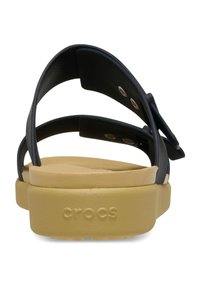 Sandalias negras con tiras y plataforma beige. Material de plástico suave y detalle de hebilla ajustable. Logo "CROCS" visible en la suela.