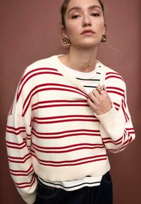 Weißer Pullover mit roten Streifen und V-Ausschnitt, getragen über einem schwarz-weiß gestreiften Hemd. Verfügt über eine lockere Passform und eine gerippte Struktur.