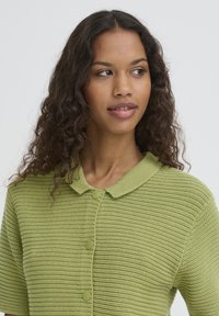 Cardigan vert en maille avec un motif texturé, manches courtes et col arrondi. Comprend des boutons ronds verts le long du devant.
