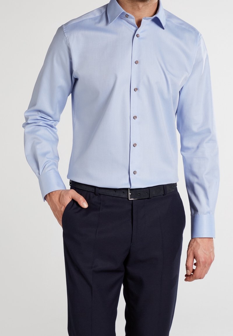 Homme portant une chemise bleu clair à boutons, rentrée dans un pantalon bleu marine foncé avec une ceinture noire, une main dans la poche, sur un fond uni.