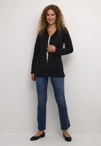 Svart texturerad cardigan med volangkanter, två framfickor och matchande knappar, ihop med blå tightsbyxor och svarta ballerinaskor.